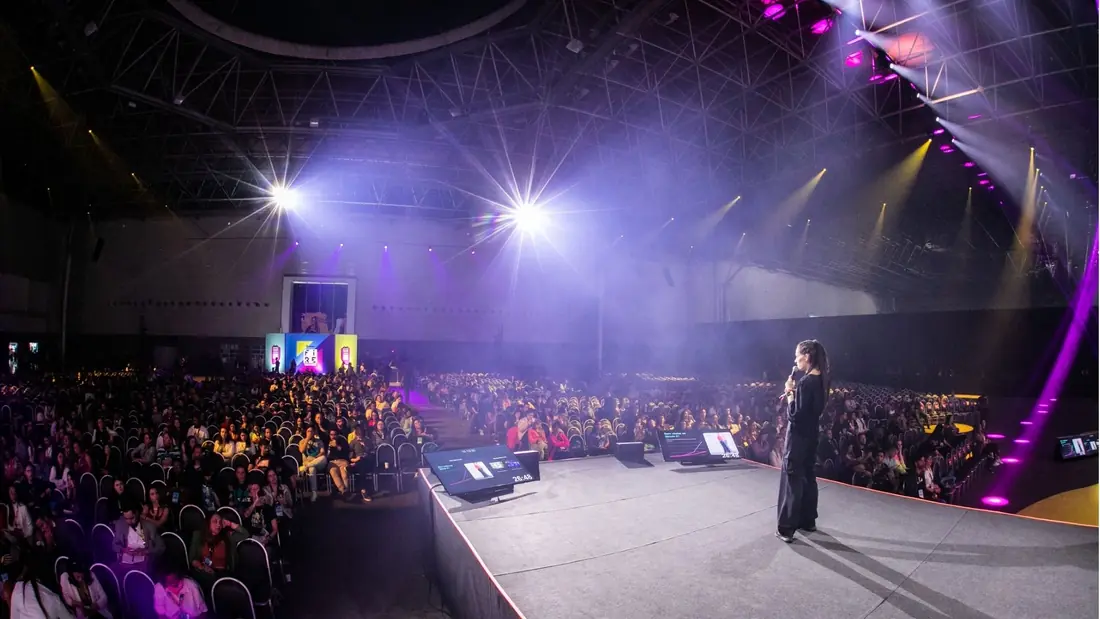Monica Magalhaes ao microfone na beira do palco, diante de um auditório lotado sob luz roxa
